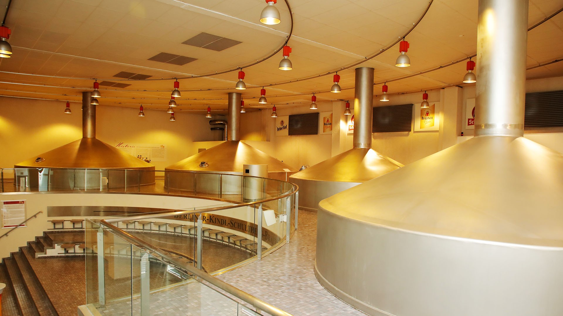 Große Braukessel aus Metall in einer industriellen Halle mit Treppen und roten Lampen.