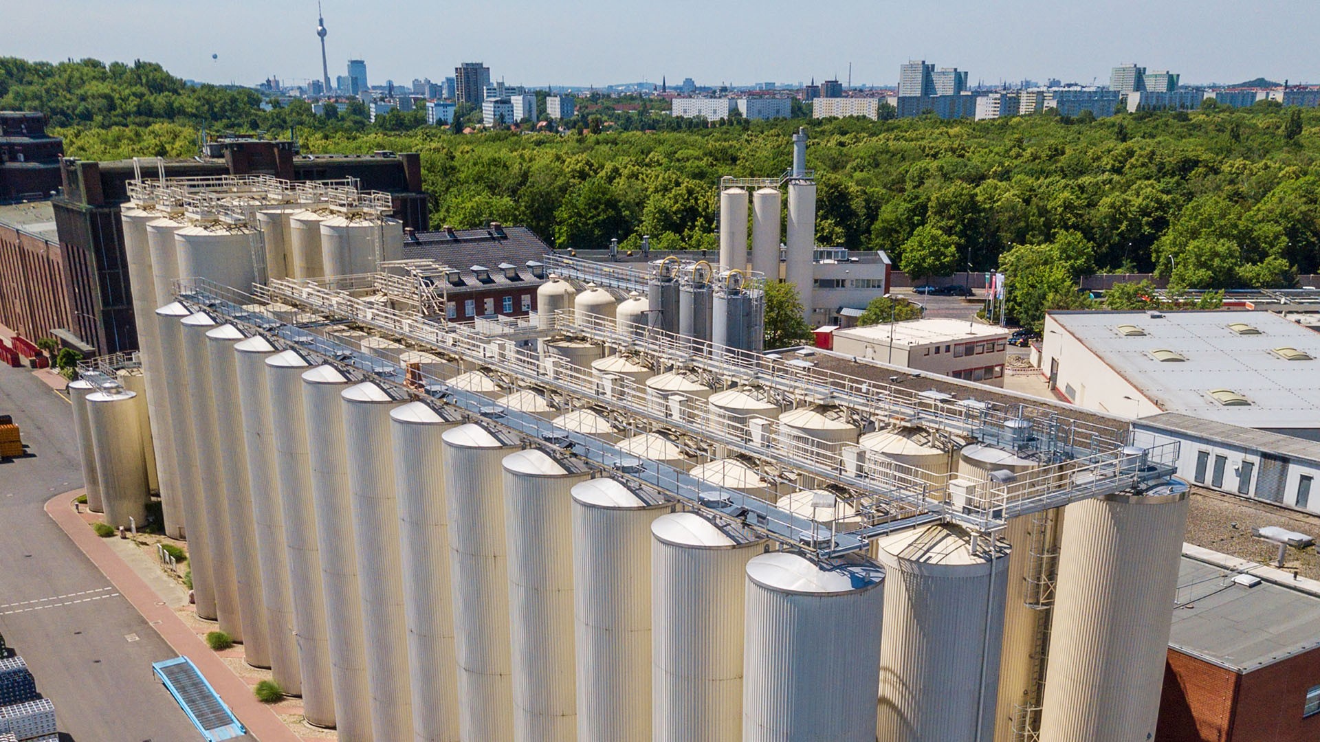 Mehrere große Silos und Tanks mit Industriebauten auf einem Fabrikgelände, umgeben von Bäumen.