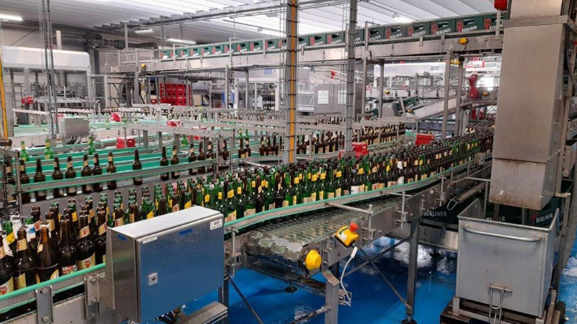 Eine große Industriehalle mit Fließbändern voller grüner Bierflaschen. Überall sind Maschinen und Förderstrukturen zu sehen.
