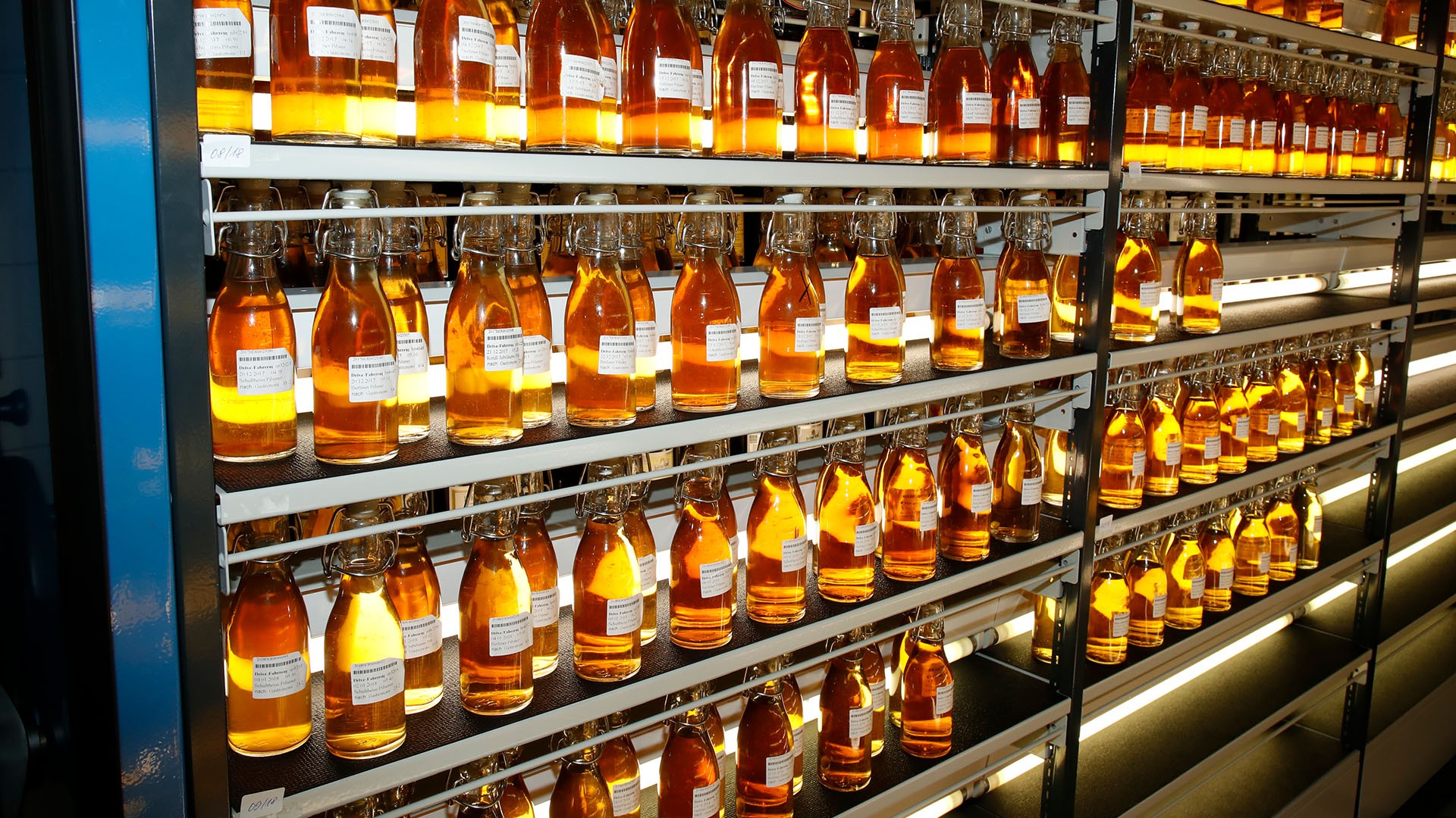 Zahlreiche Flaschen mit gelber Flüssigkeit sind auf Regalen in einer Fabrik aufgestellt. Jede Flasche trägt ein Etikett.