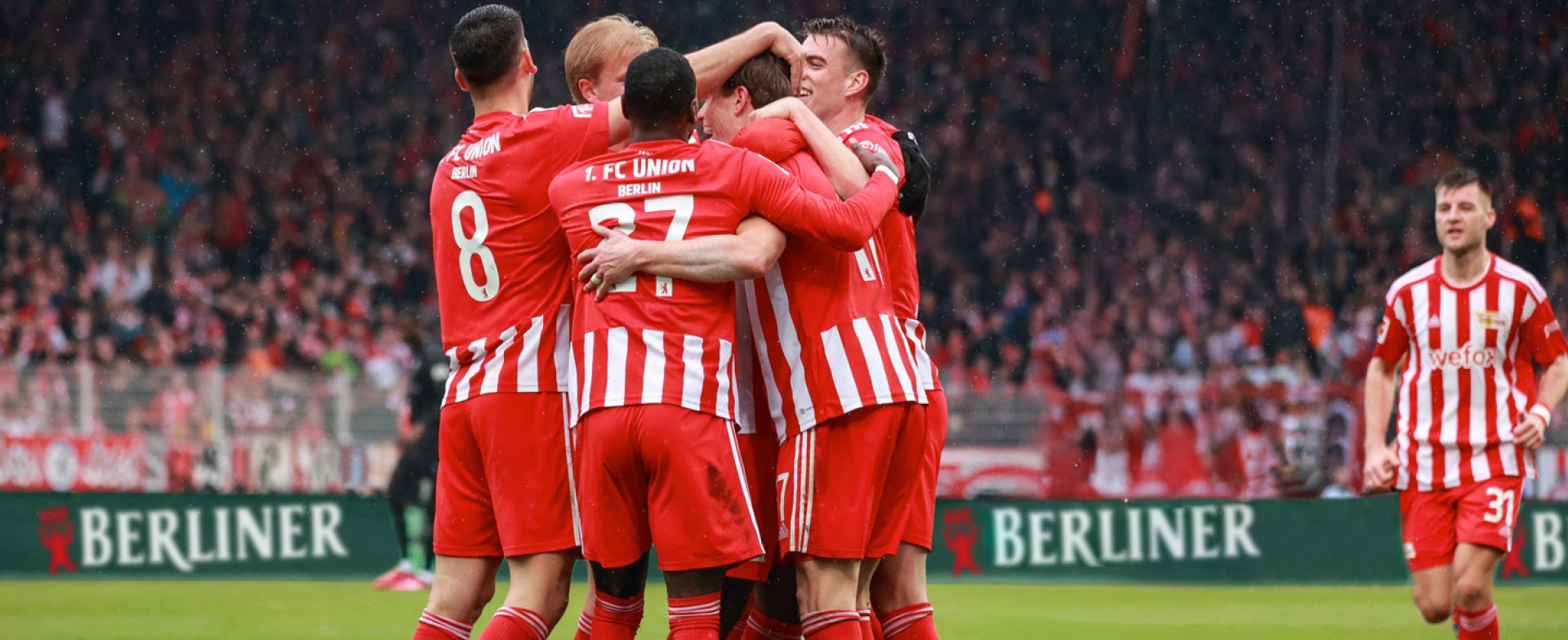 Fünf Fußballspieler von Union Berlin umarmen sich auf dem Spielfeld und feiern ein geschossenes Tor.