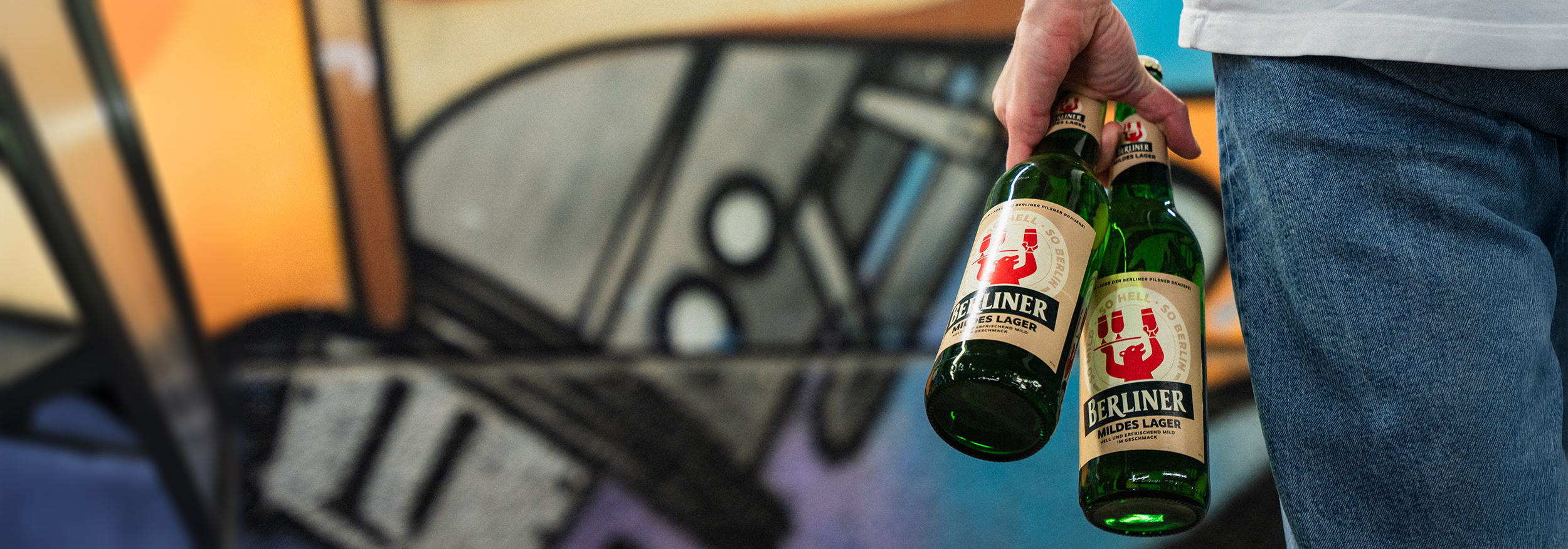 Eine Person mit zwei Flaschen Berliner Mildes Lager in der Hand steht vor einer Grafitti bemalten Wand.
