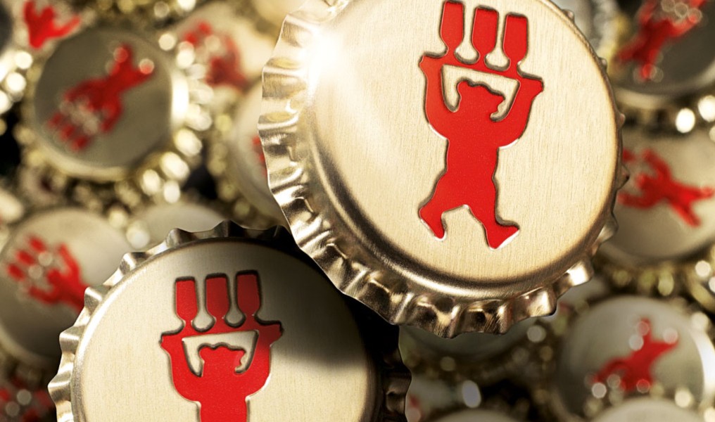 Mehrere Kronkorken sind eng beieinander angeordnet, einige im Fokus, andere unscharf. Jeder Deckel trägt ein rotes Symbol in Form einer tanzenden Figur.
