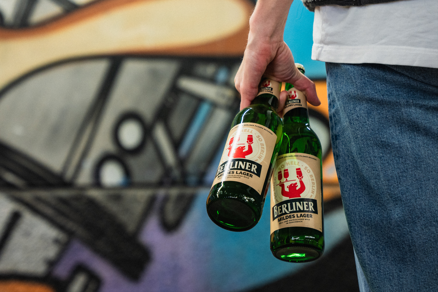 Eine Person mit zwei Flaschen Berliner Mildes Lager in der Hand steht vor einer Grafitti bemalten Wand.